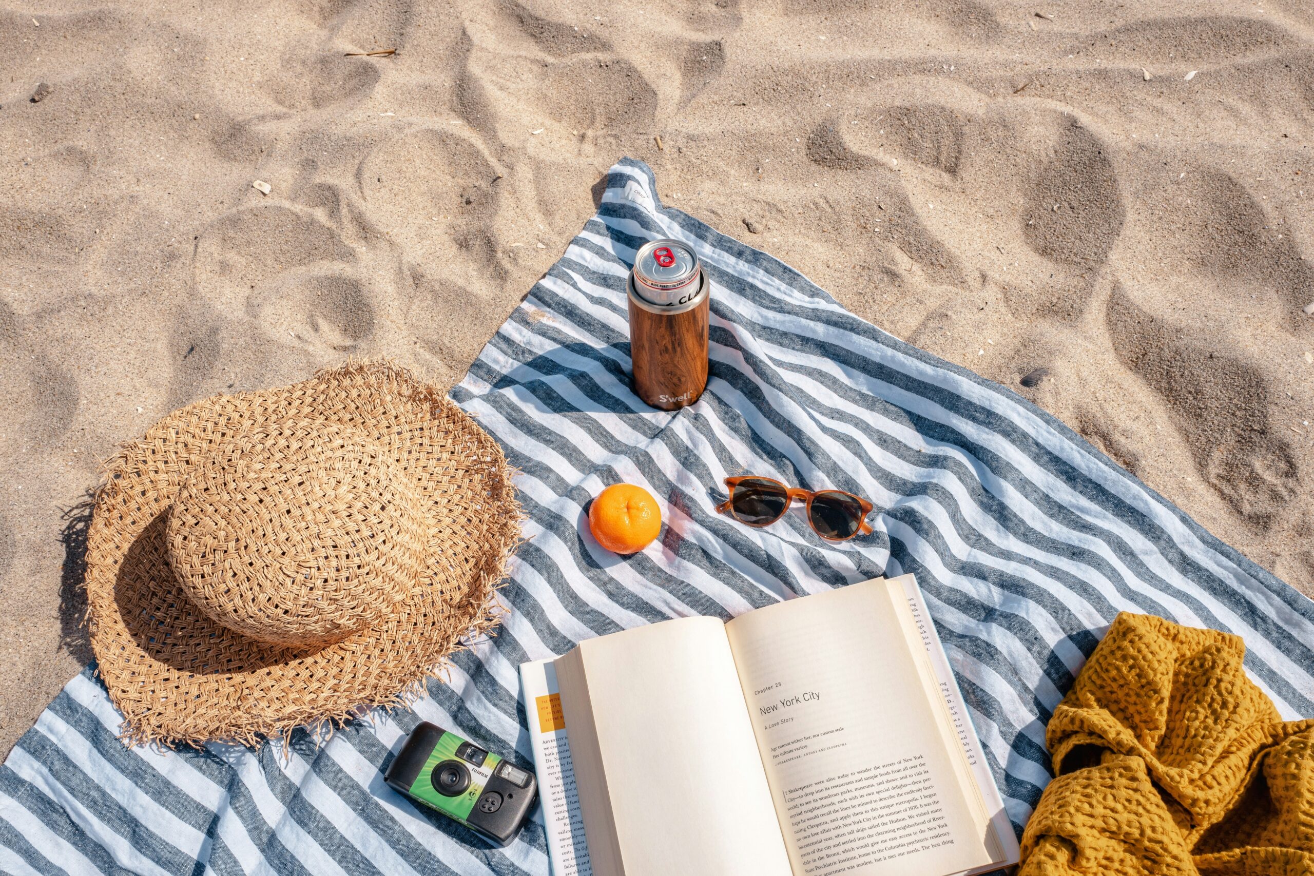 Verschieden Strandutensilien auf einem Badetuch: Ein Strohhut, Kamera, Sonnenbrille, Buch, eine Dose, ein gestrickter Überwurf und eine Orange